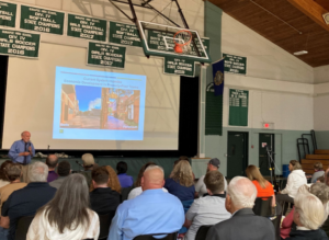 An audience of people in a gymnasium watch a speaker and slideshow