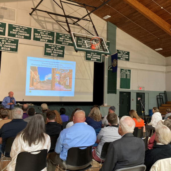 An audience of people in a gymnasium watch a speaker and slideshow